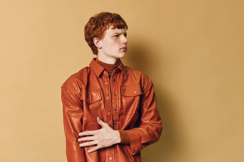 Young man with red hair wearing orange leather jacket posing and looking sideways against plain beige background in studio portrait with thoughtful expression and modern style.