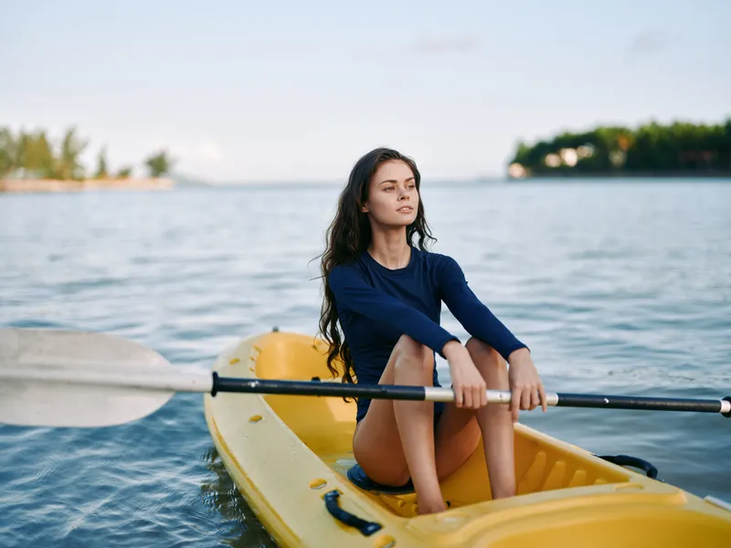 Young woman sits in a yellow kayak on calm blue water, wearing a navy long sleeve top, gazing into the distance, capturing a serene outdoor paddling moment.