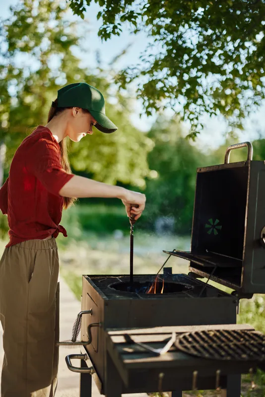 Young woman grilling outdoors in a green park, showcasing summer cooking skills with a cheerful expression, wearing casual clothing and a cap