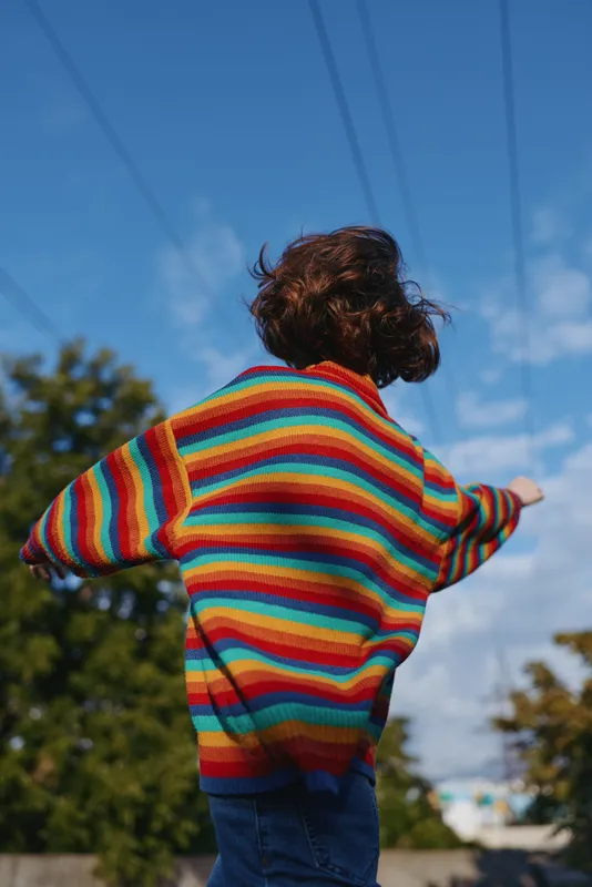 Child in rainbow sweater back view with arms outstretched outdoors under blue sky, playful childhood scene in casual clothing and curly hair, motion and joyful freedom.
