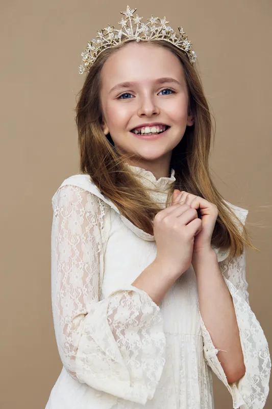 Young girl with a crown smiling in a light dress against a beige background, conveying joy and innocence, ideal for children s themes and celebrations