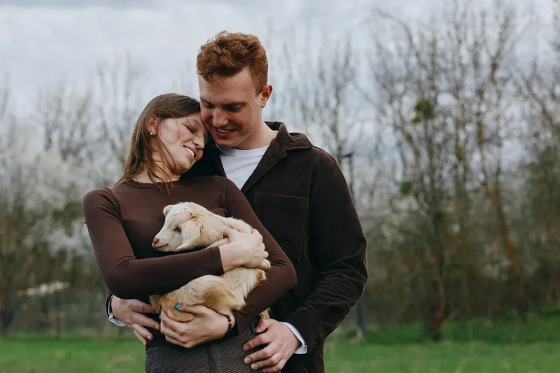 Joyful couple outdoors share a tender embrace with a light puppy, smiling and cozy in fall attire, natural park background, candid affectionate moment, warm lifestyle scene, everyday happiness