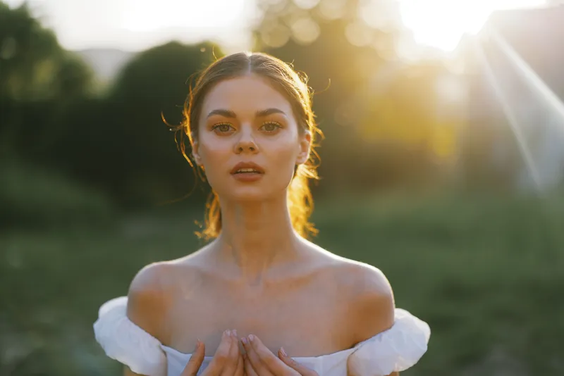 Ethereal young woman in a white dress with bare shoulders, standing in a sunlit green field, conveying serenity and natural beauty in a dreamy atmosphere