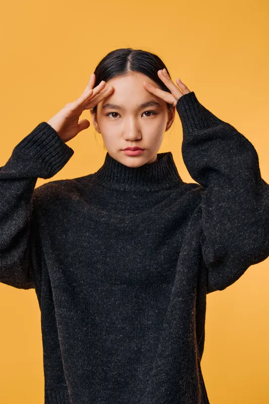 Young woman with long black hair wearing dark sweater standing in front of orange background looking at camera with thoughtful expression and hands touching temples.