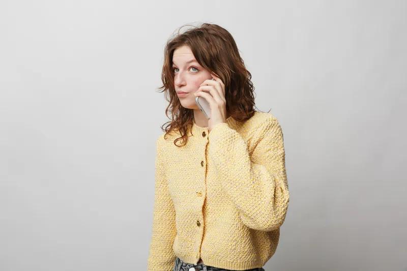 Young woman with curly hair wearing yellow knitted sweater talks on smartphone. Isolated on light background. Concept of communication, technology, casual style, and modern lifestyle.