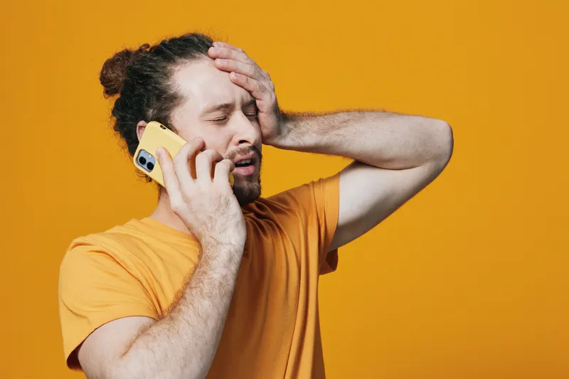 A frustrated young man talking on the phone, showing emotion against a vibrant orange background, wearing a casual yellow t shirt, perfect for stress related concepts