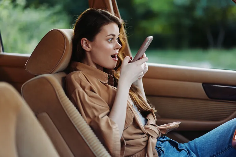 Young woman surprised by a message on her phone while sitting in a car, wearing a casual beige shirt, captured in a vibrant outdoor setting.