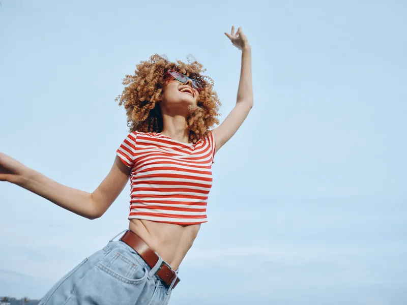 Young dancer in a striped crop top and jeans pivots and leaps under a clear blue sky, embodying radiant energy, freedom, and carefree movement in a sunlit outdoor moment today, vivid.