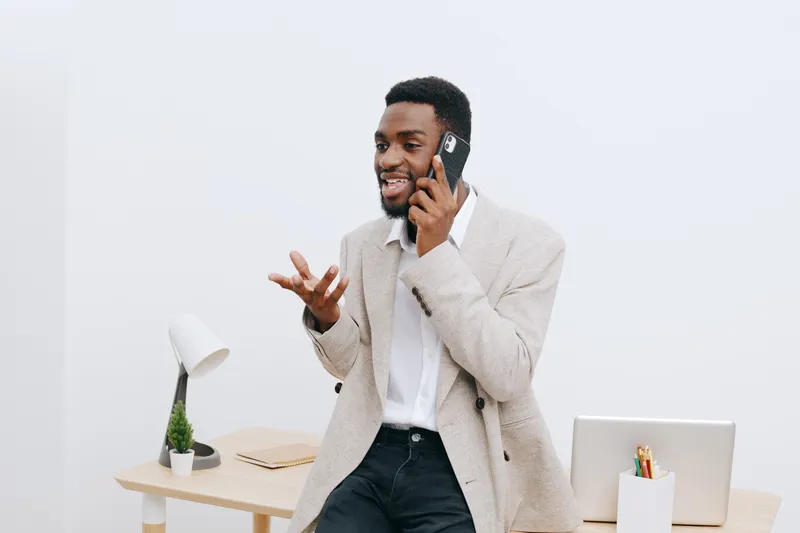 Smiling young African man wearing a light-colored blazer talking on his smartphone in a modern office setting with minimal decor and a white background. People lifestyle concept