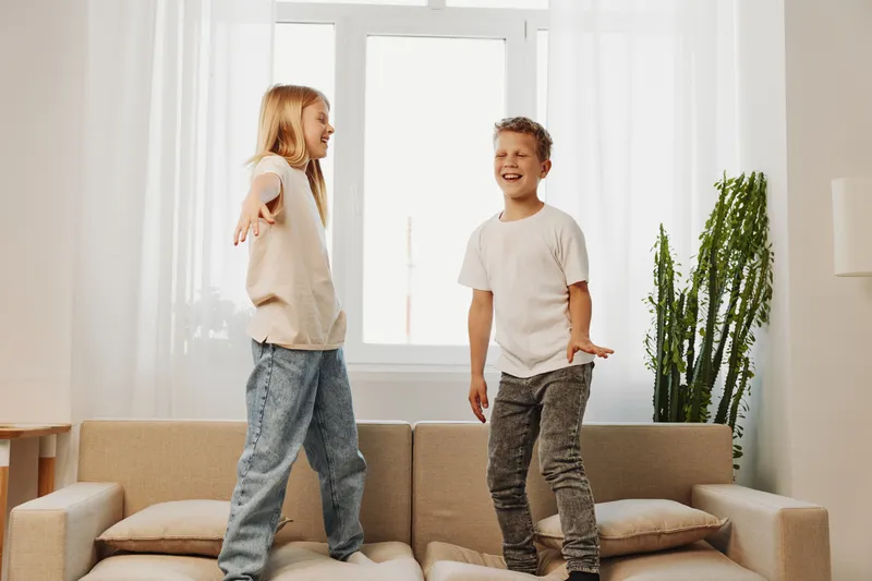 Two happy children playing on a beige couch in a bright living room with natural light. They are smiling and enjoying carefree moments of fun and friendship indoors.