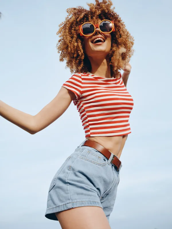 Joyful young woman dancing outdoors in a striped crop top and denim shorts, sunlit sky behind, carefree expression, vibrant summer vibe and energetic movement.