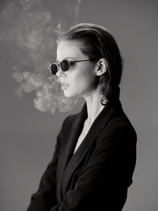 Black and white portrait of a stylish woman in sunglasses exhaling smoke, dressed in dark clothing, with wet hair and a confident expression