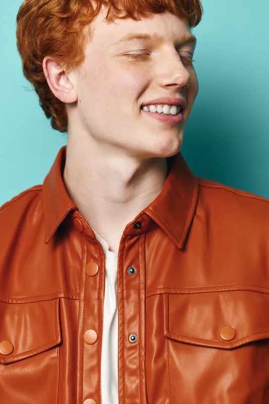 Young man with red hair wearing brown leather jacket and white shirt smiling looking sideways against blue background for casual fashion portrait style.
