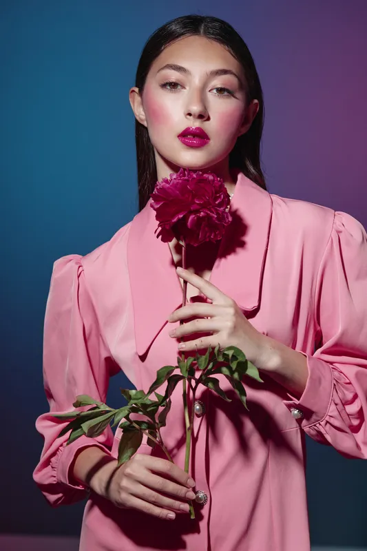 Young woman with dark hair in a pink dress holds a dark pink flower with green leaves against a colorful background. Fashion beauty creative portrait concept.