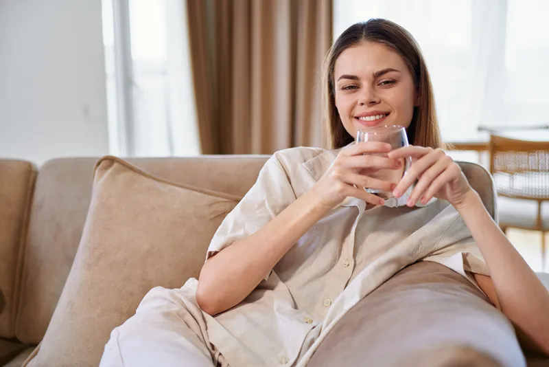 Woman with smile holding a glass of water on a sofa at home, relaxation and comfort, casual lifestyle portrait of drink and cozy livingroom scene with natural light and calm.