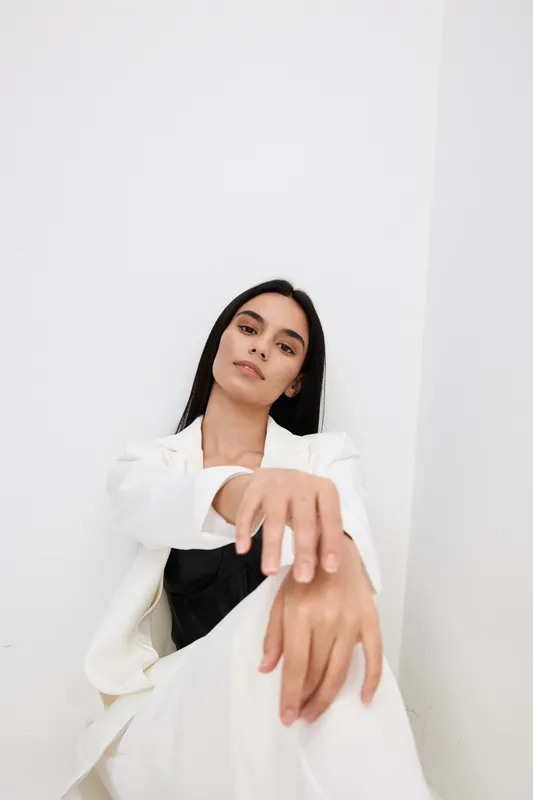 Young woman with long dark hair wearing white jacket and black top, sitting relaxed against white wall, showcasing calm expression and minimalist style.