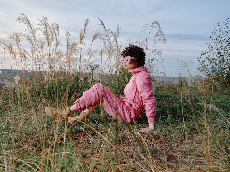 Outdoor fashion scene with a person in a pink jumpsuit sitting in tall grass on a hillside, relaxed pose, natural landscape and calm mood.