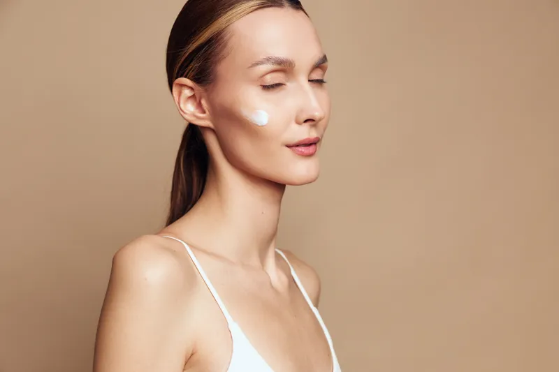 A young woman with closed eyes applies moisturizer cream on her cheek in a beige studio. The concept represents skincare, beauty routine, hydration, and self-care for healthy skin.