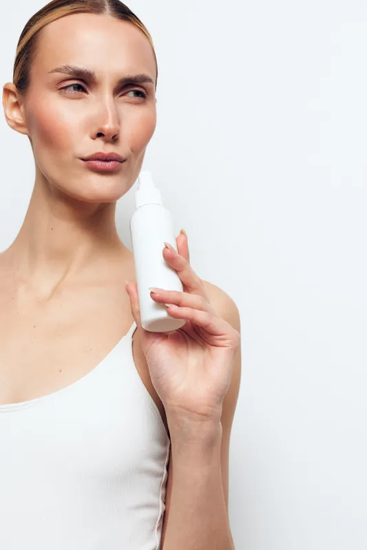 A young woman in a white top holds a white spray bottle near her face, looking to the side with a thoughtful expression. The background is plain white, emphasizing skincare concept.