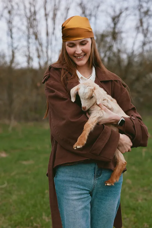Cheerful woman outdoors, holding a goat kid with a gentle smile, warm sunlight, casual attire, relaxed pose, natural colors, grassy field, rustic setting, candid moment, affectionate mood