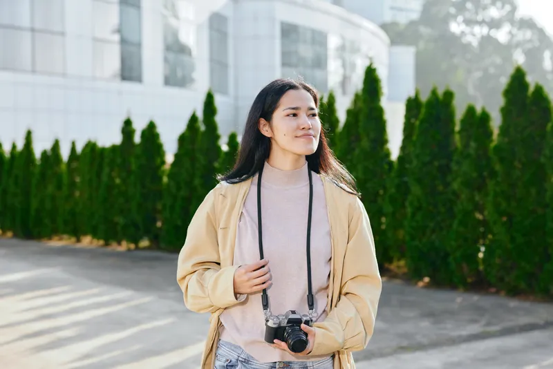 Casual photographer with camera in hand stands outdoors near a modern building, wearing a beige jacket and soft sweater, capturing daylight scenes with confident focus and poised posture