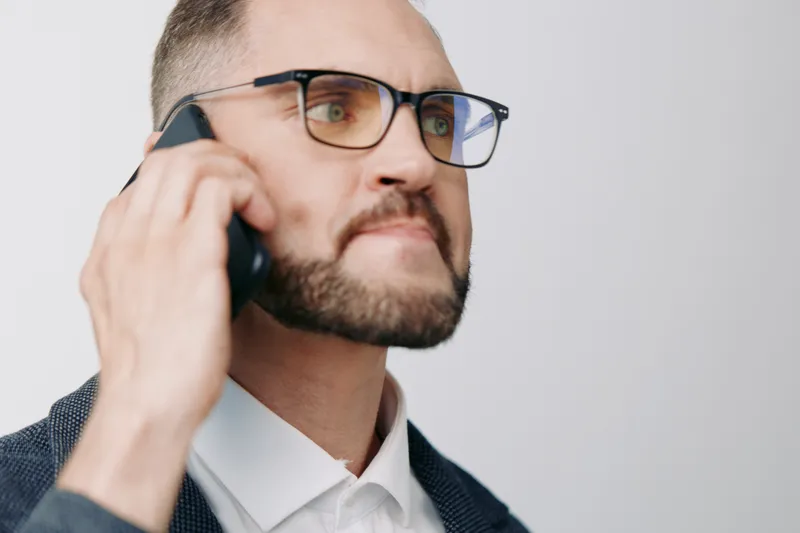 A business professional man on a call stands against a single colored background, showing confidence and focus. His composed expression and smart attire convey leadership in a corporate setting.