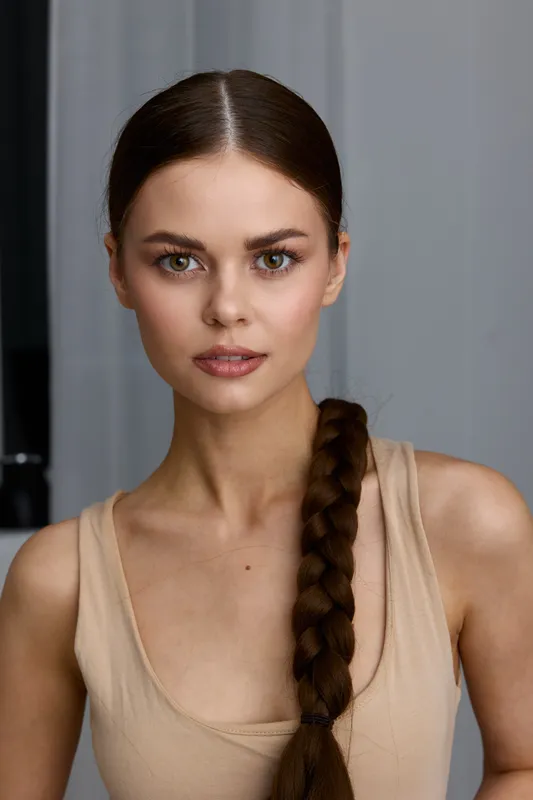 young woman with long braided hair wearing a beige top looks confidently at camera with neutral gray background and natural makeup, close-up portrait.