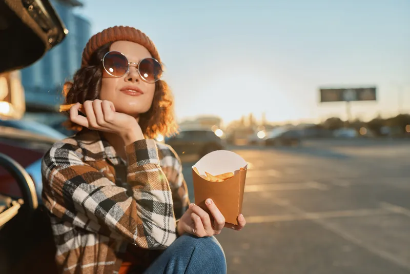 Woman with fries and sunglasses in a beanie by a car on a street at golden hour, candid portrait capturing mindful living, authenticity and emotional storytelling in urban lifestyle.
