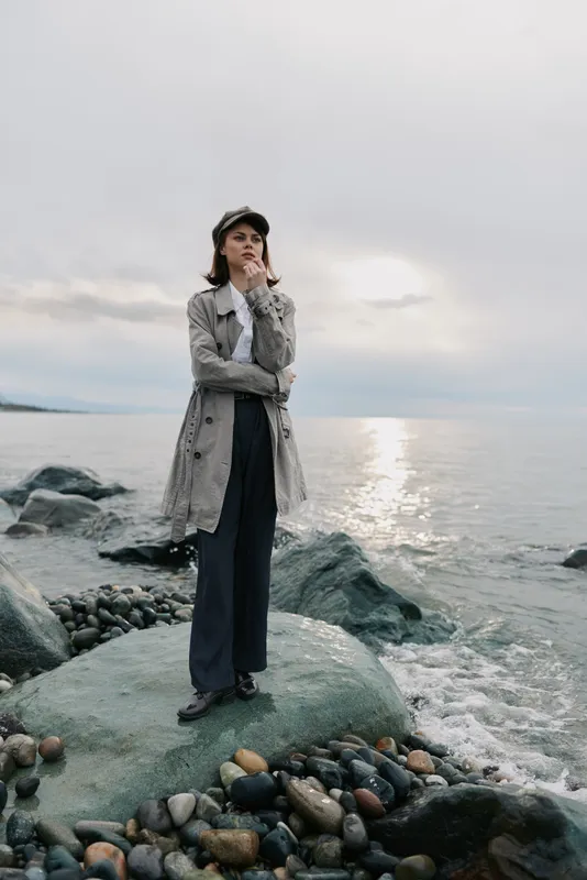 Young woman in long coat and hat stands on rocky beach near calm sea during cloudy day. Reflective water and moody sky create peaceful atmosphere for outdoor fashion portrait.