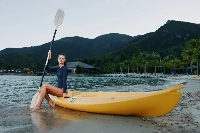 Young woman sits on a yellow kayak at the shore, holding a paddle, ready for water adventure, with calm waves, sandy beach, and lush mountains in the background.