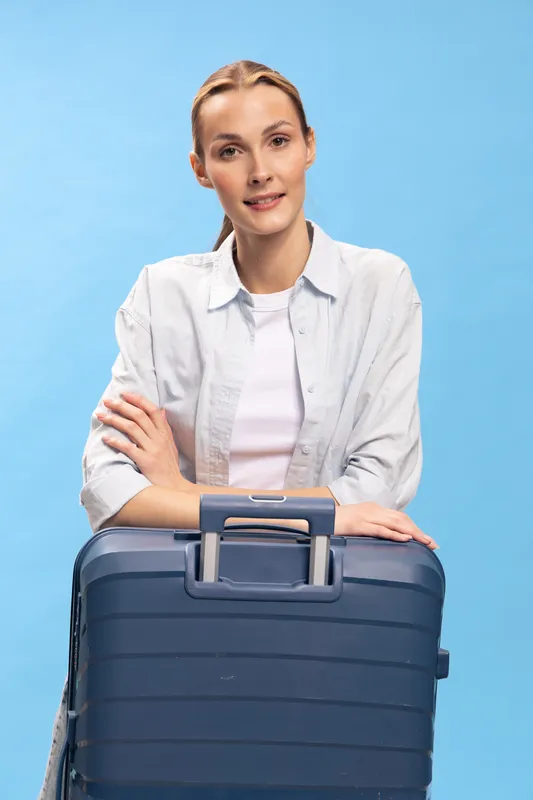 Young woman with folded arms leans on blue suitcase, smiling gently. Casual outfit and light background create relaxed mood. Concept for travel, vacation, and preparation.