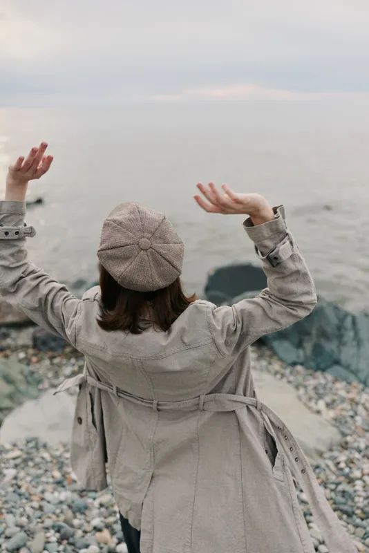 Woman in plaid coat and hat raises hands while standing on rocky beach near calm sea under cloudy sky. Autumn fashion and peaceful nature concept with casual vintage style outfit.