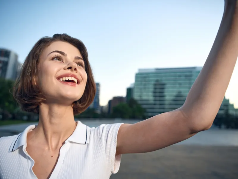 A cheerful young woman takes a selfie in a sunny urban park beside a river, smiling brightly as modern buildings rise in the background, conveying optimism, confidence, and a casual, upbeat lifestyle