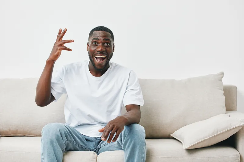 Cheerful young Black man sitting on a sofa, smiling broadly, wearing a casual white t shirt and jeans, expressing joy in a modern, bright living room