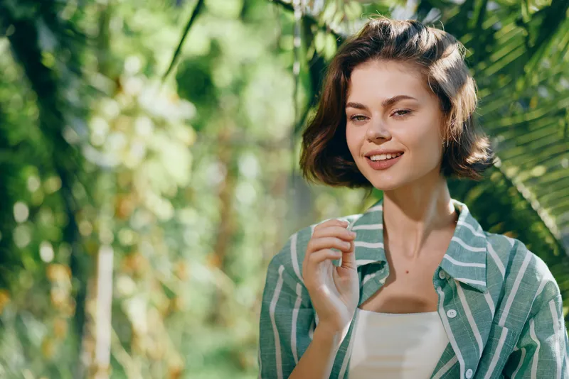 A smiling young woman stands in a sunlit garden, wearing a striped shirt, capturing a relaxed, natural moment.