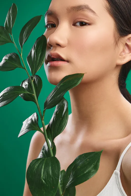 asian woman in a studio portrait with a leafy plant close to her face against a green backdrop, soft lighting, calm mood, and minimal elegant styling.