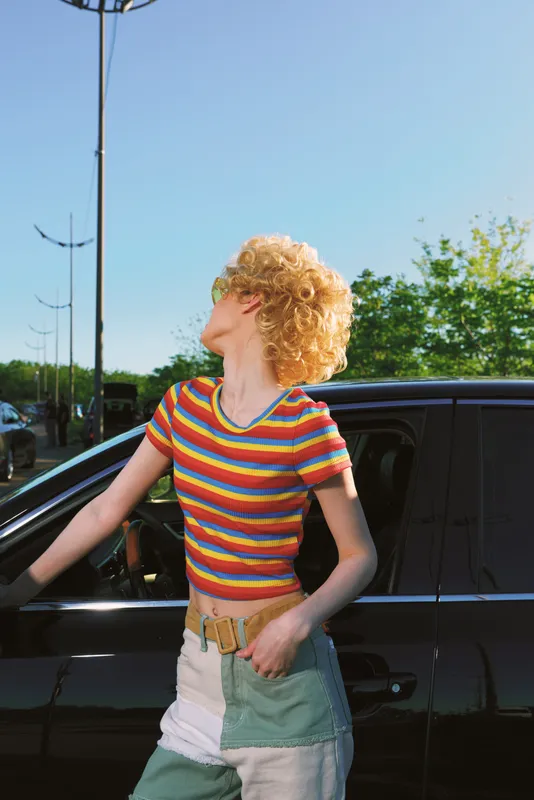 Vibrant woman outdoors wearing a rainbow striped shirt, curly hair, candid smile, sunlight glow, casual pose, car side scene, bright colors, natural setting, relaxed lifestyle