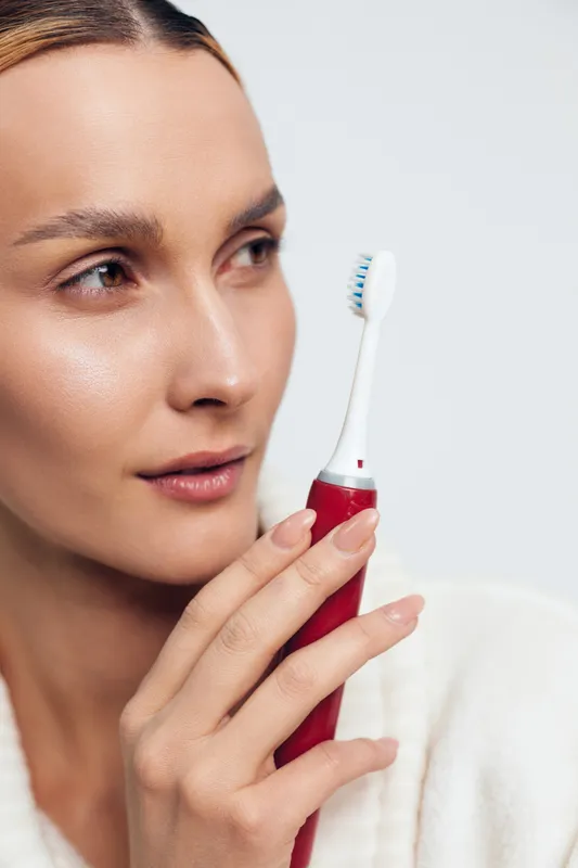 Young woman holding electric toothbrush near face, closeup portrait, clean skin, focused expression, white background, oral hygiene, personal care routine.