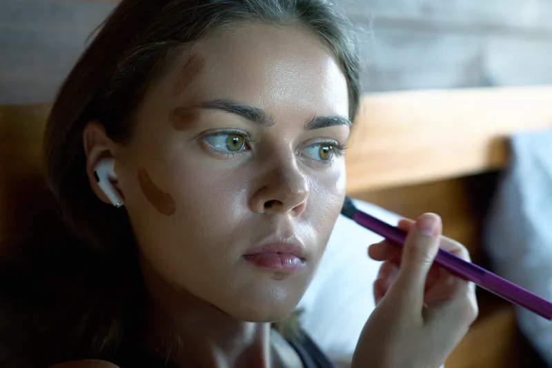Woman applying makeup indoors, closeup portrait with soft natural light, skincare routine vibe, cosmetics focus, relaxed mood, candid lifestyle moment, gentle colors