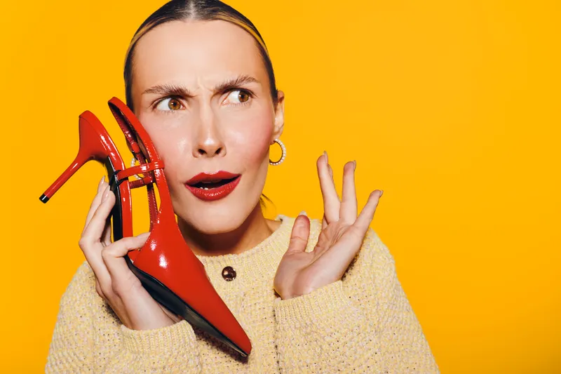 Confused woman in yellow sweater holds red high heel shoe near ear on bright orange background. Concept for fashion, style, footwear, and unusual communication.