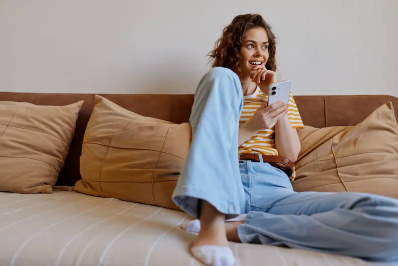 Relaxed woman indoors on a sofa, smiling while texting on a smartphone, striped top, blue pants, warm natural light, cozy home ambiance, casual lifestyle moment captured with soft shadows