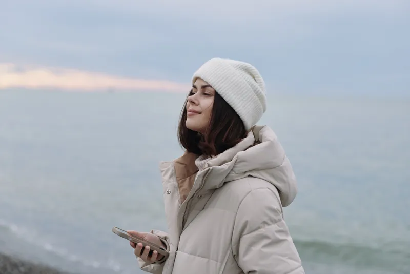 woman winter coat beanie outdoor by the sea, calm portrait as she holds a smartphone against a serene coastal breeze.