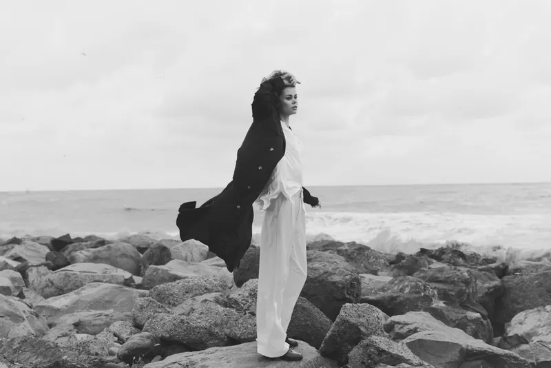 Monochrome scene with a standing woman on rocky shore, wind sweeping cloak behind, vast sea horizon, contemplative mood, dramatic coastal composition