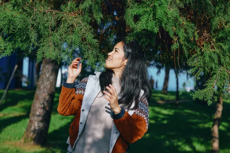 Woman enjoying mist from a sprinkler under pine trees, outdoors in a park, wearing a cozy sweater, playful moment of hydration and fresh air, joyful expression in sunlight