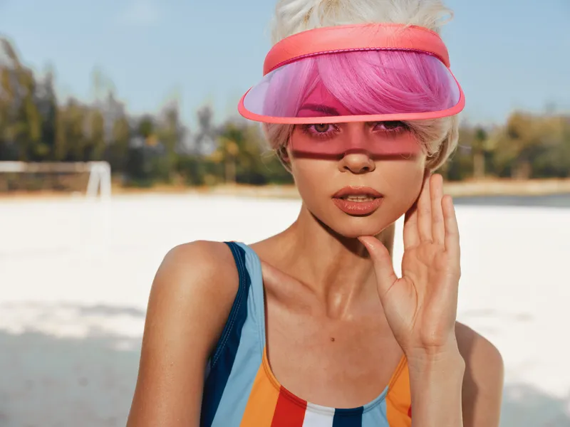Portrait of a stylish woman at a sunny beach wearing a colorful striped swimsuit and a pink visor, sunlit skin, relaxed pose, and a chic summer mood.