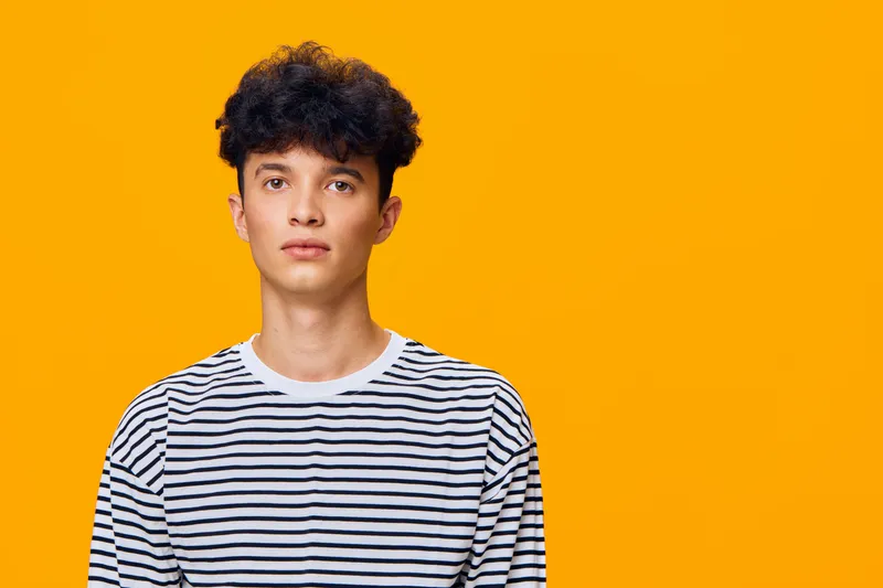 Young man with curly hair wearing a black and white striped shirt stands against a bright orange background, exhibiting a calm and neutral expression.