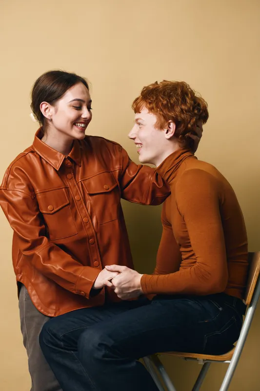 Couple sitting and holding hands, smiling and looking at each other with affection and warmth in casual clothes against beige background. Young adults sharing moment.