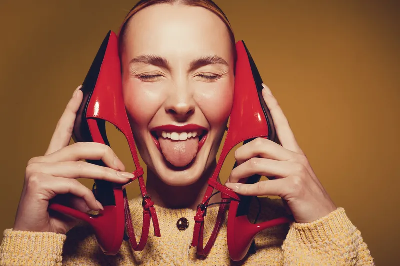 Happy young woman with closed eyes holds red high heels near face and shows tongue. Warm yellow background and cozy sweater create playful and joyful mood in studio portrait.