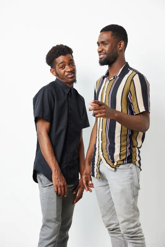 Two African American men share a moment of laughter, wearing stylish outfits. Their expressions reflect friendship and joy in a light, neutral setting.