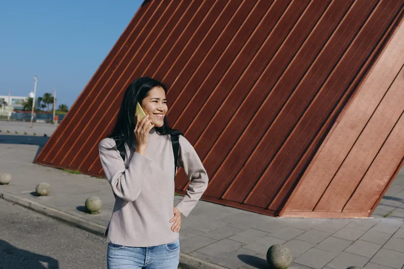 Woman on phone outdoors beside a bold angular wall, casual clothes, smiling as she chats, urban architecture backdrop, street scene, sunny day, modern vibe, everyday moment